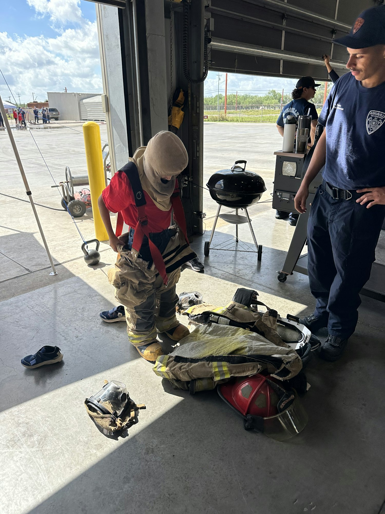 People in a garage with firefighting gear on the floor and a grill nearby.