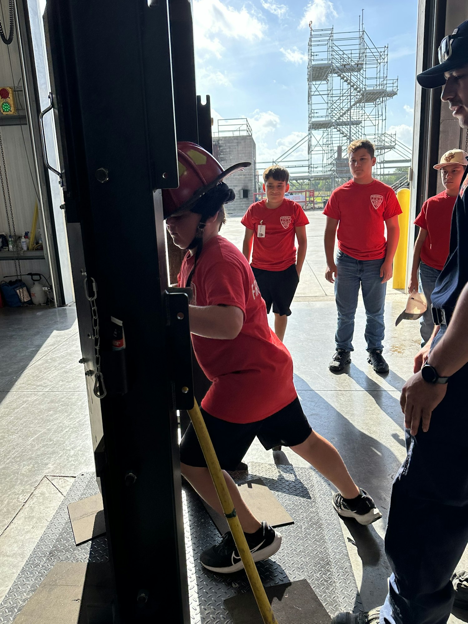 A group of people in red shirts and helmets are gathered, possibly training or at an event, with a ladder and structures in the background.