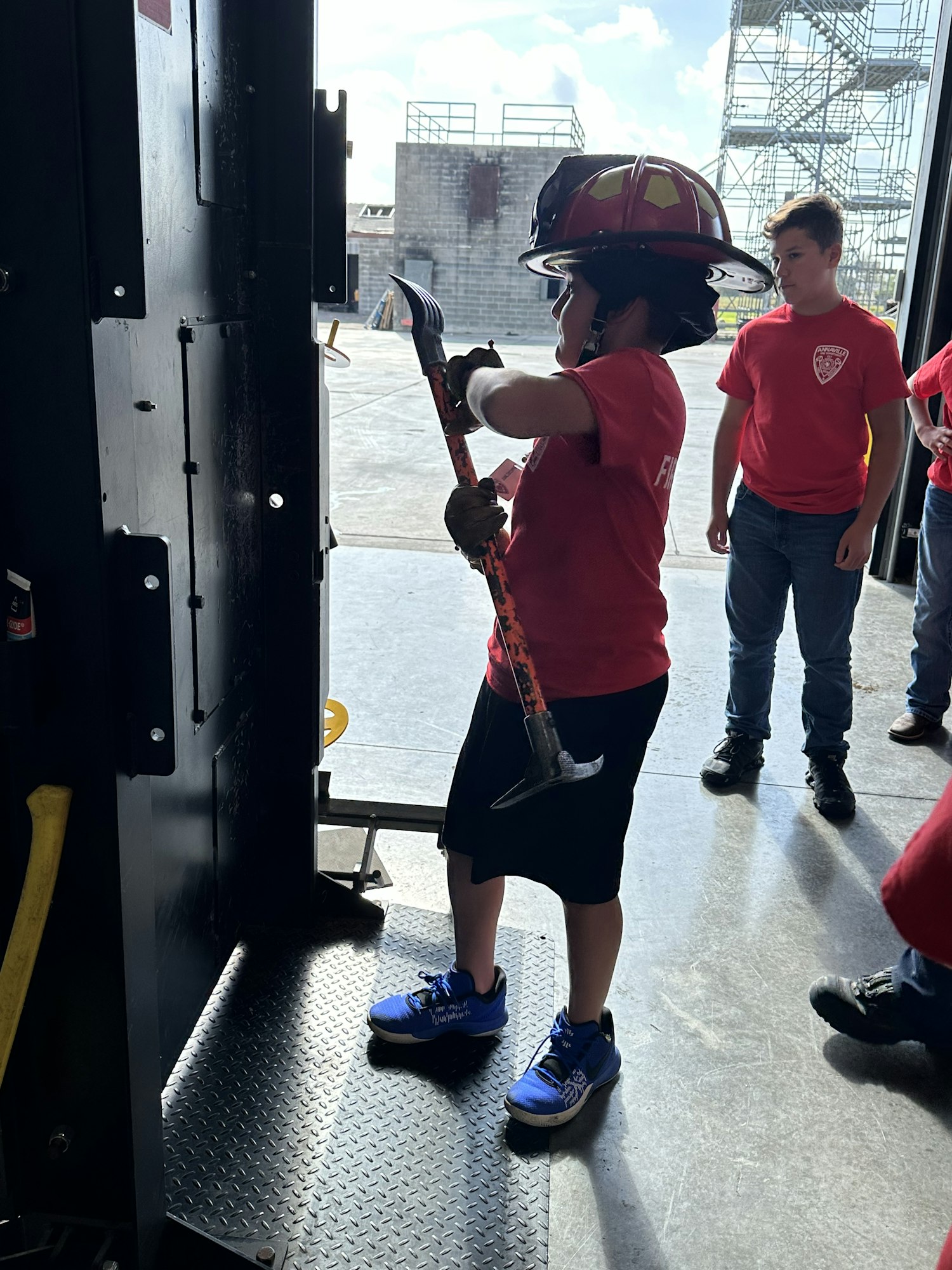 A child in a firefighter helmet holds a tool, surrounded by others wearing red.