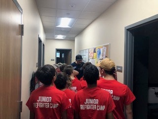 A group of people in red "Junior Firefighter Camp" shirts standing in a hallway, facing a person in a dark outfit.