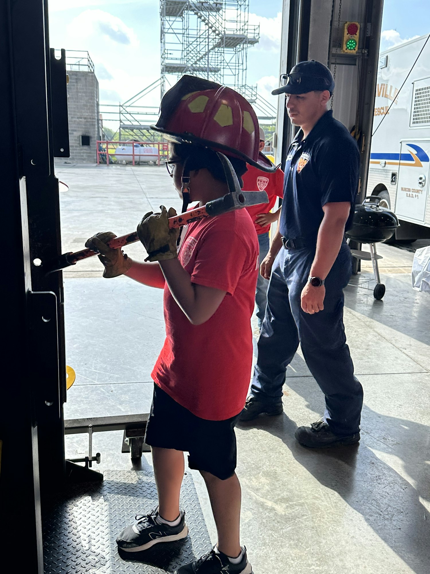 A child wearing a firefighter helmet and gloves is holding a tool, with an adult standing nearby in a garage.