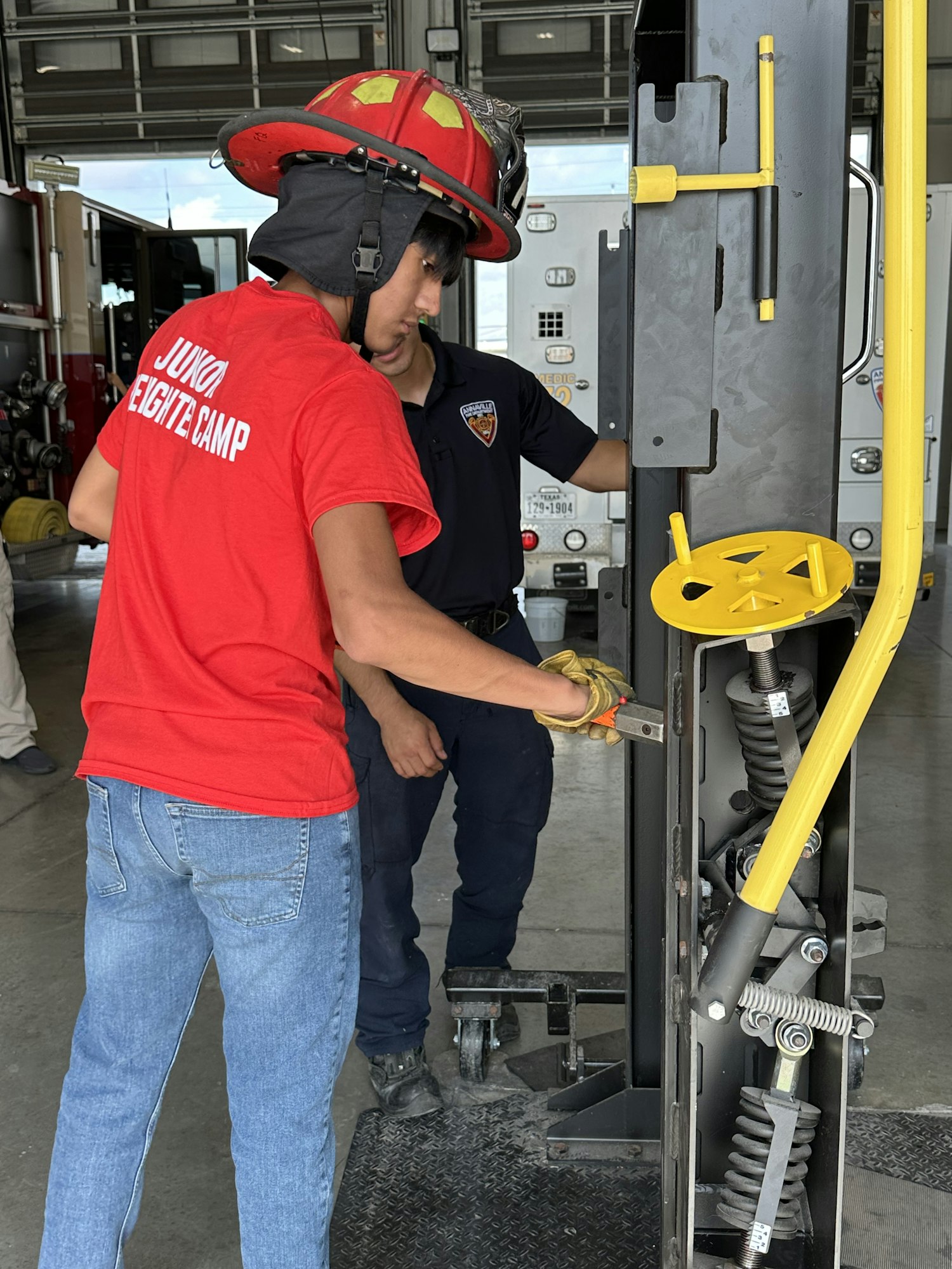 Two people, one in a "JUNIOR FIREFIGHTER CAMP" shirt and helmet, work on a mechanical equipment.