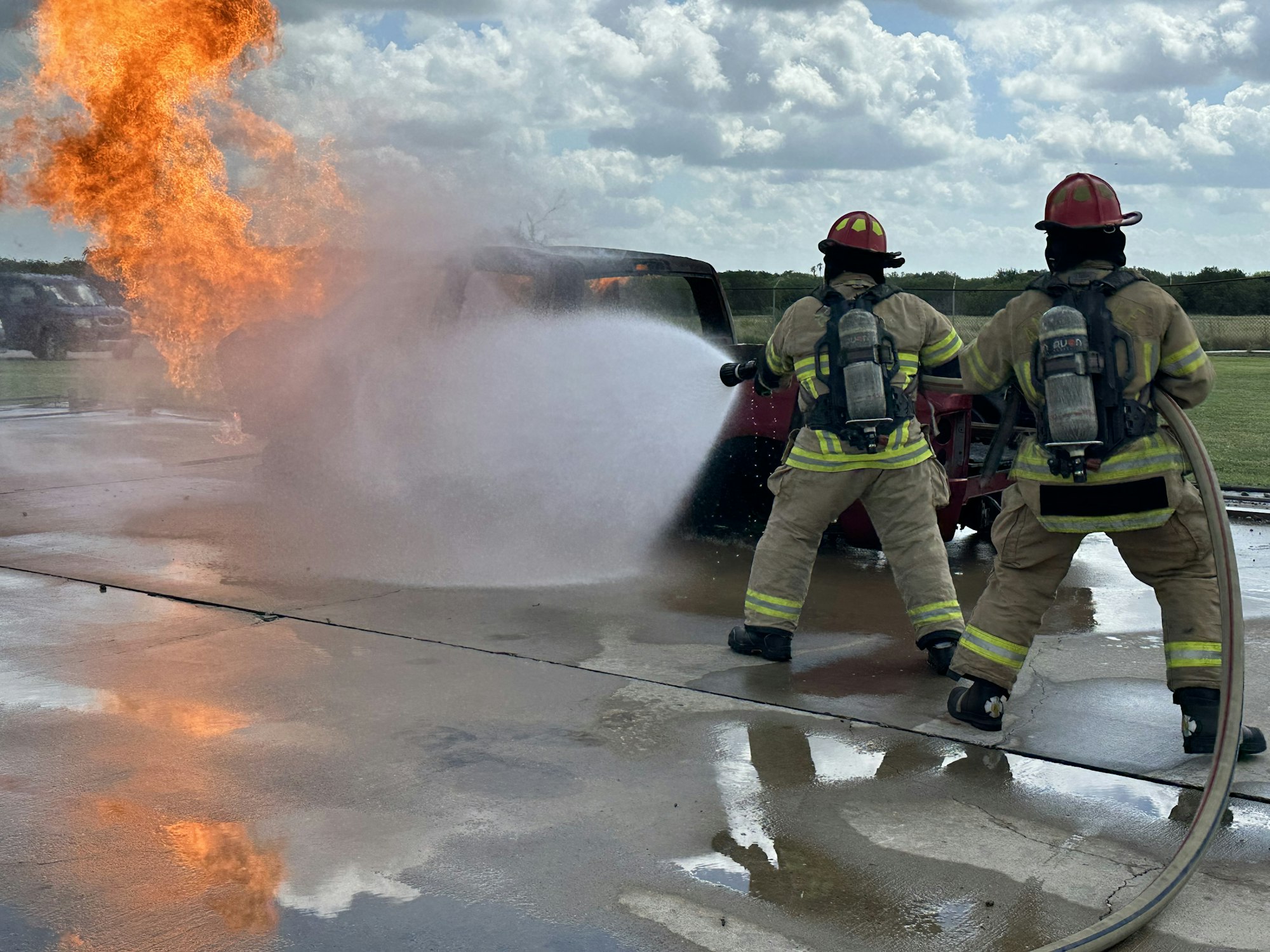 Firefighters extinguishing flames from a car, using a hose with water, wearing protective gear and helmets.