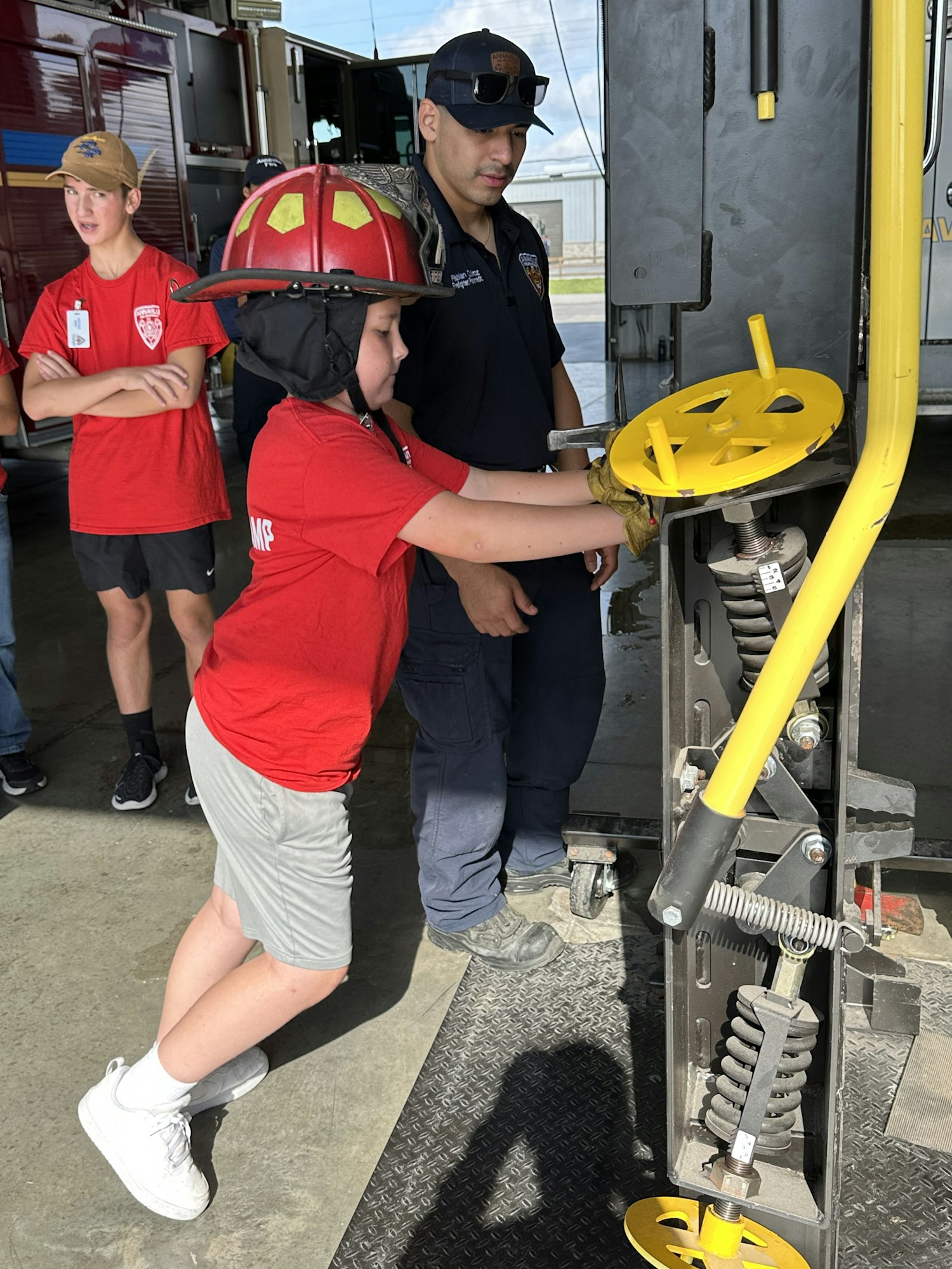 A person in a red shirt and helmet operates a yellow valve, observed by others in a workshop setting.