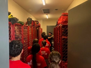 Individuals in red shirts are in a narrow room with red lockers and gear bags.