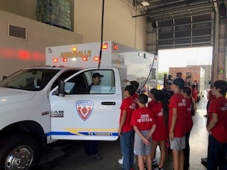 A group of kids in red shirts stand next to an ambulance, listening to a person in the driver's seat inside a building.