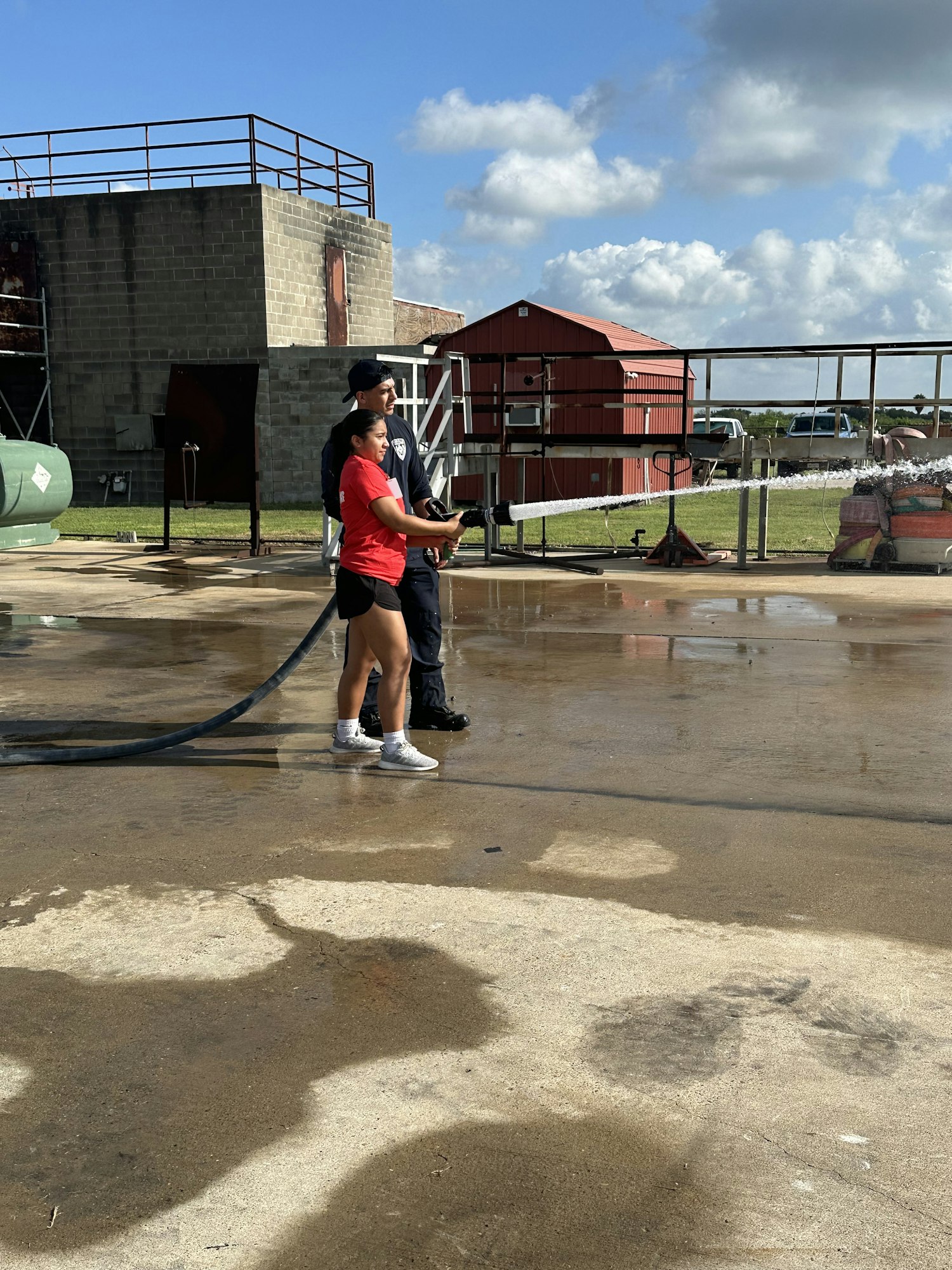 Two people using a firehose outdoors, with a building and a clear sky in the background.