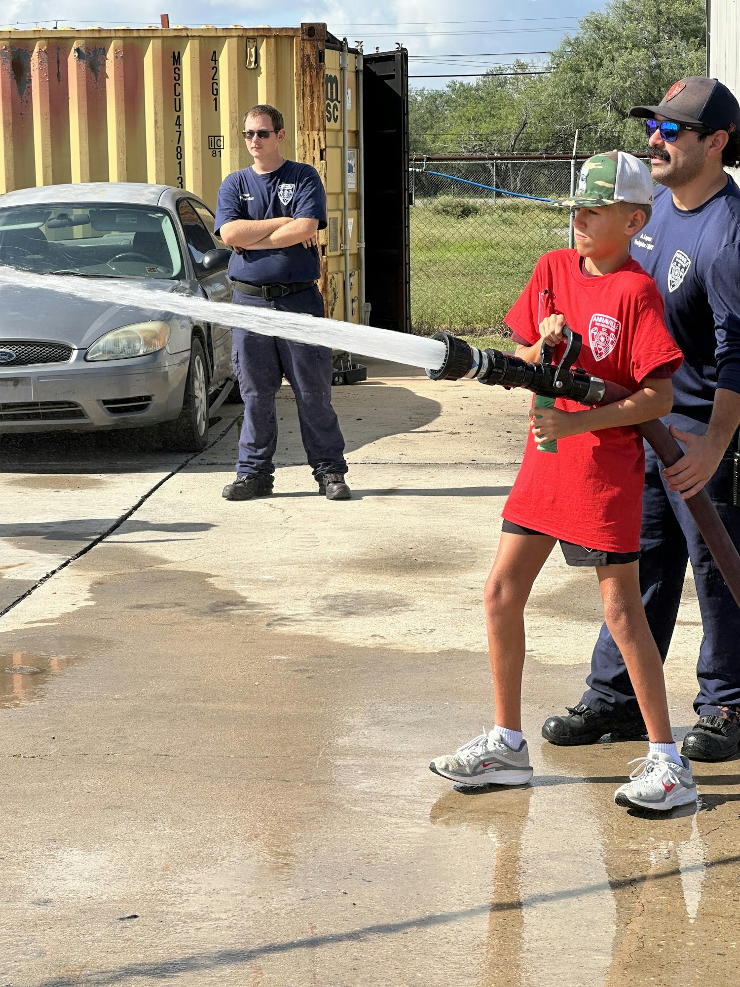 A person in a red shirt uses a fire hose, assisted by two others, near a car and shipping container outdoors.