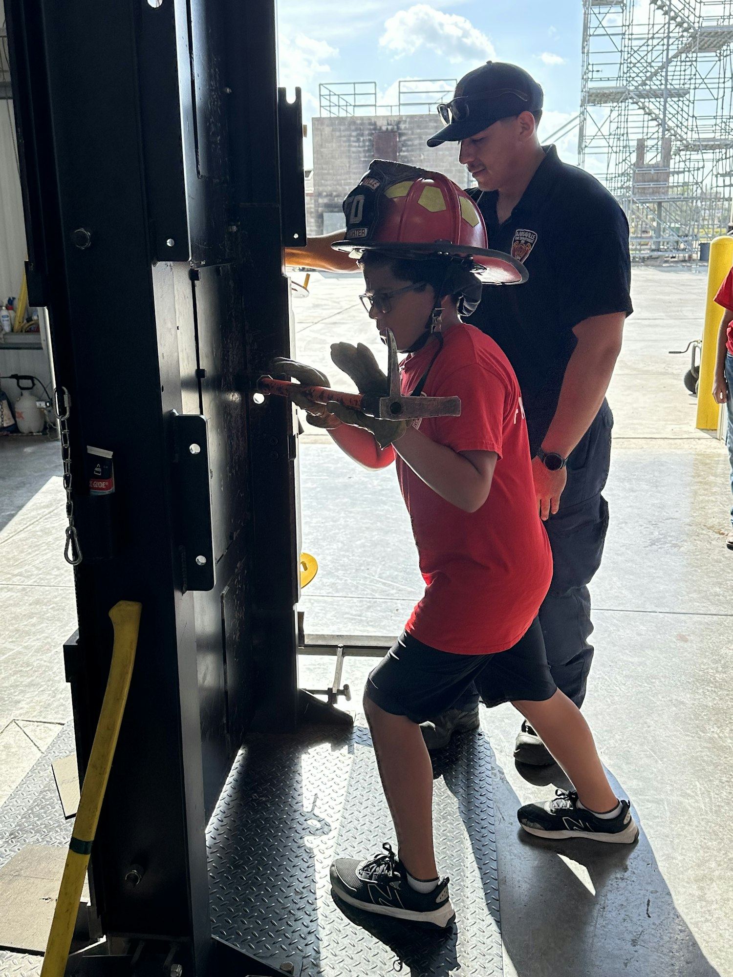 A child in firefighter gear practices using an axe under adult supervision.