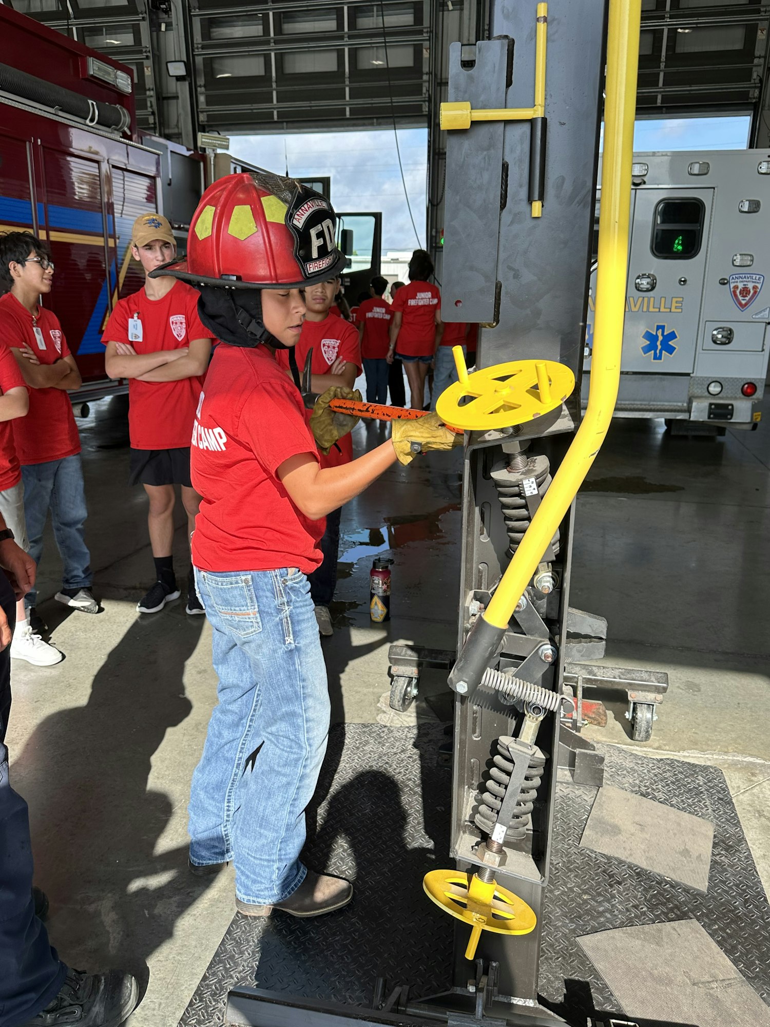 A child wearing a firefighter helmet turns a yellow wheel on a mechanical device, surrounded by others in red shirts in a fire station.