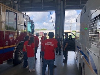A group of people in red shirts stand between a fire truck and another emergency vehicle in a garage.