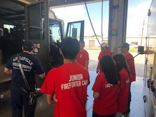 A group of people in "Junior Firefighter Camp" shirts listening to a firefighter near a fire truck inside a fire station.