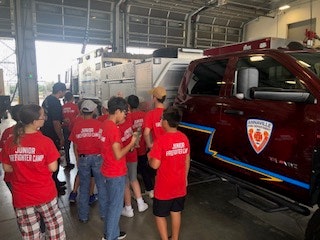 A group of kids in red "Junior Firefighter Camp" shirts gather around a fire truck in a fire station.
