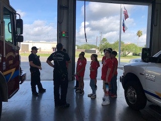 A group of people in red shirts are listening to fire personnel inside a garage with fire trucks nearby.