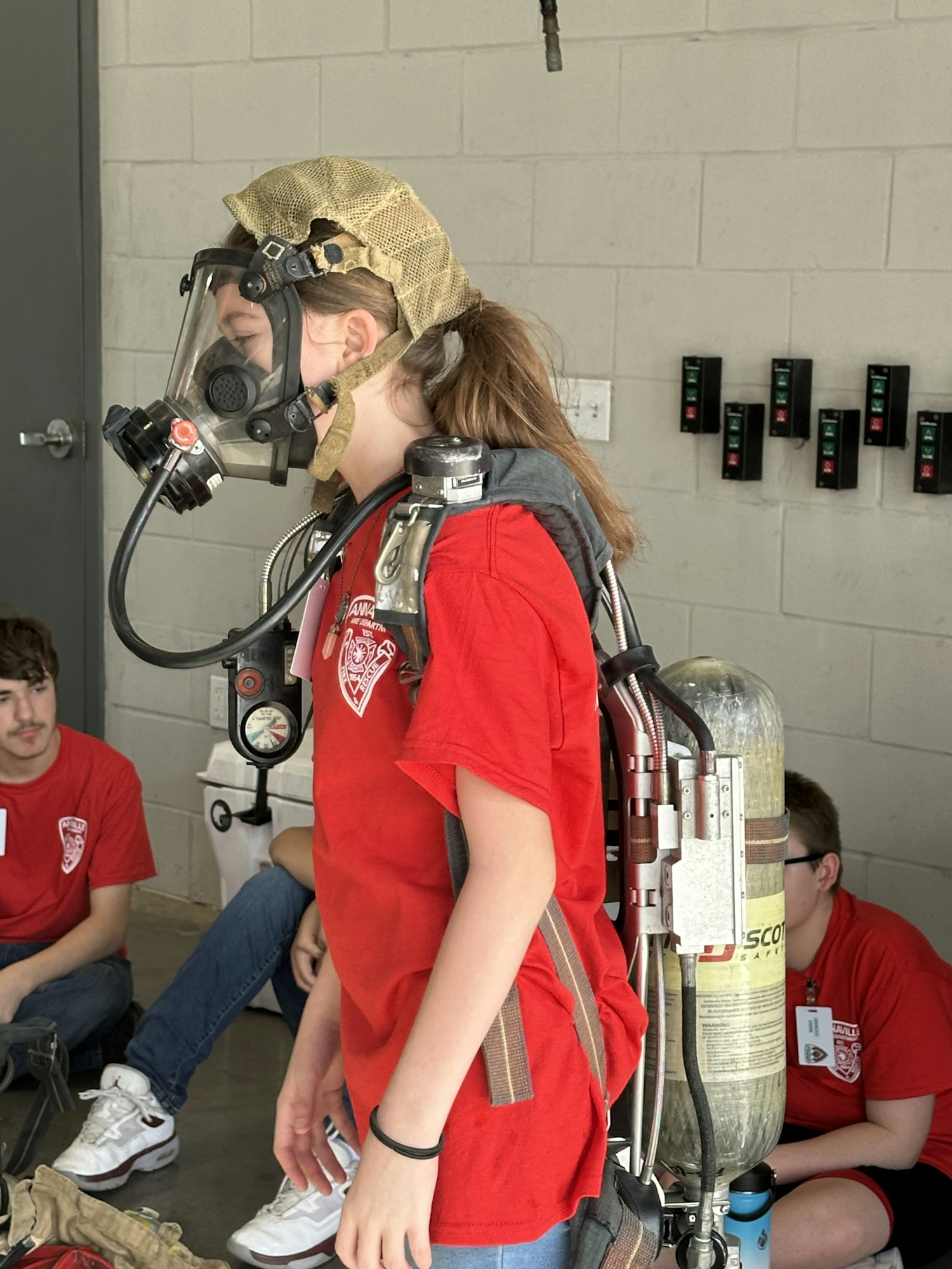 A person in a red shirt wearing a breathing apparatus, with gauges visible on a wall and another person seated in the background.