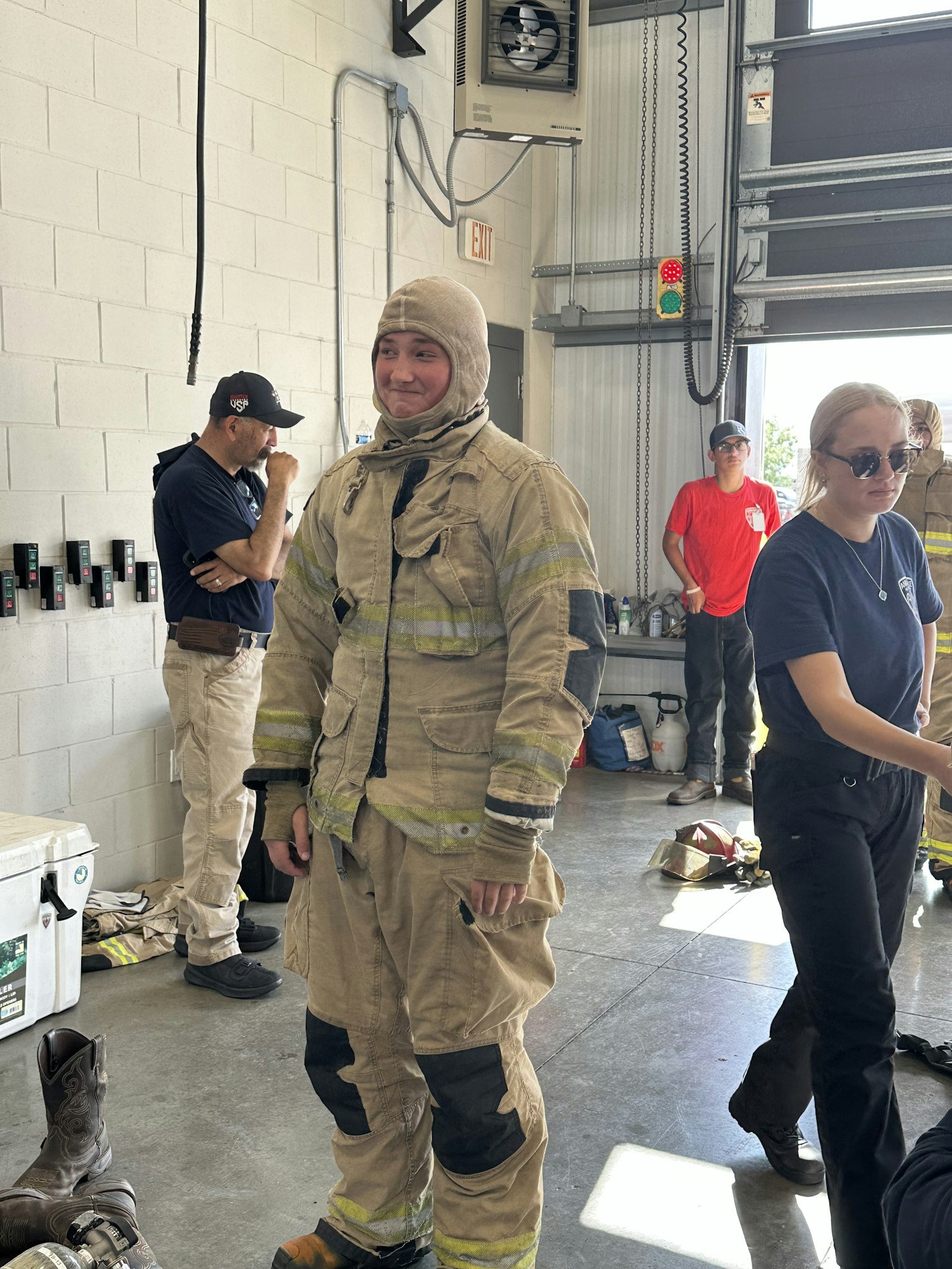 A person in firefighter gear stands indoors; others are around, one seated, some standing. Equipment and a cooler are visible nearby.