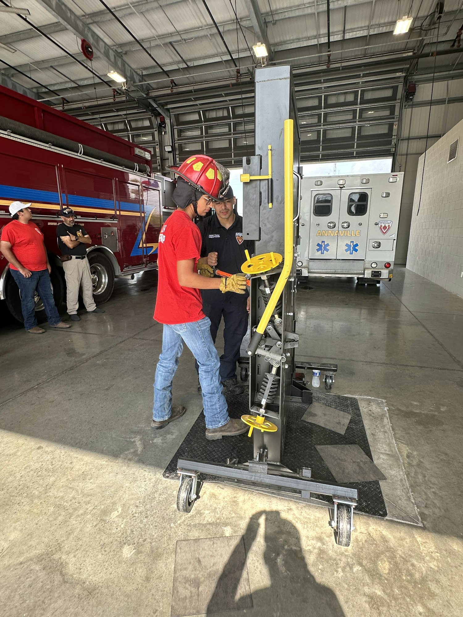People are working on machinery inside a garage with an ambulance and fire truck in the background.