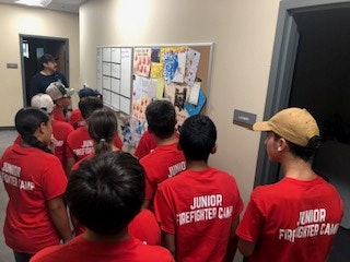 Group of kids in red "Junior Firefighter Camp" shirts looking at a wall with art and papers.