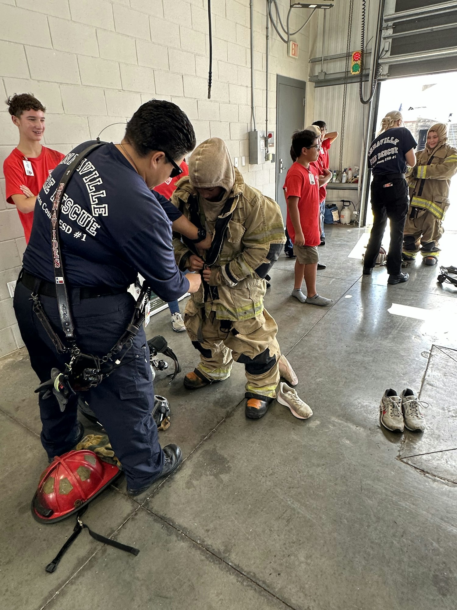 People in firefighter gear are training in a garage; others in red shirts observe or assist.