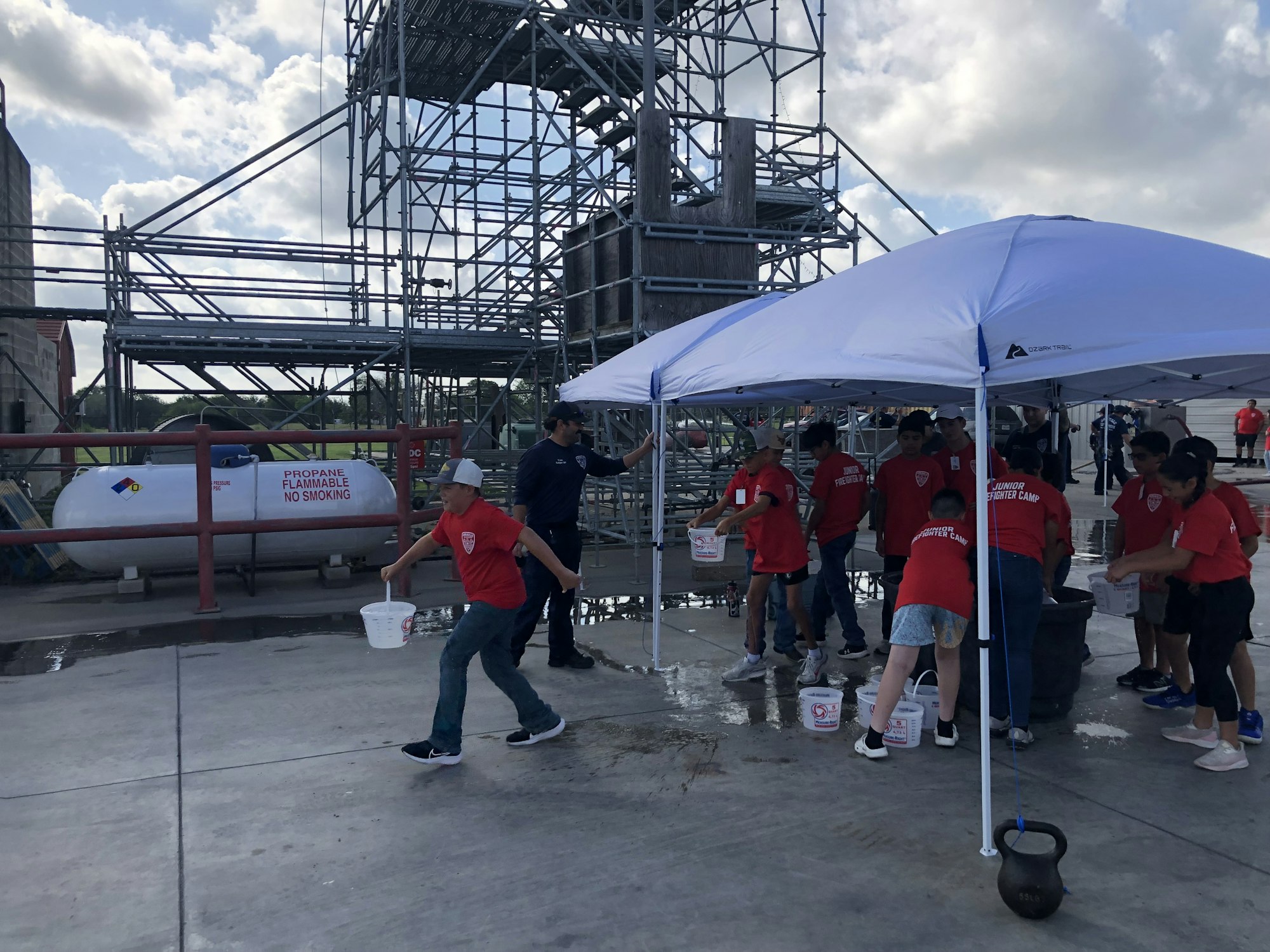 Group of kids in red shirts under a canopy, engaged in a water activity near a propane tank and scaffolding.