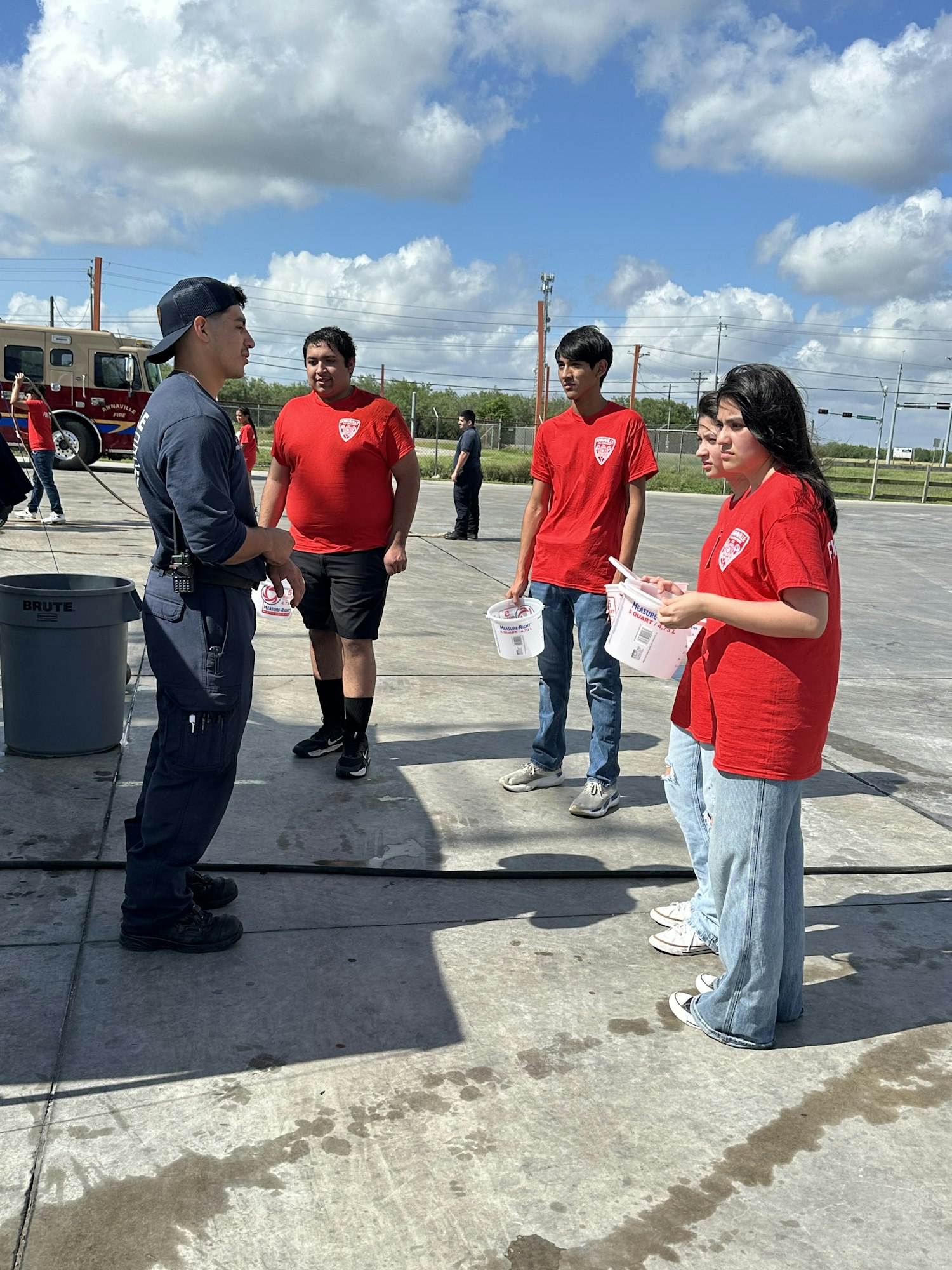 People in red shirts and a person in a dark uniform stand outdoors, holding papers, near a fire truck.