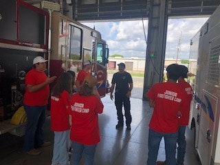 A group of people in "Junior Firefighter Camp" shirts stand near fire trucks, listening to a firefighter.