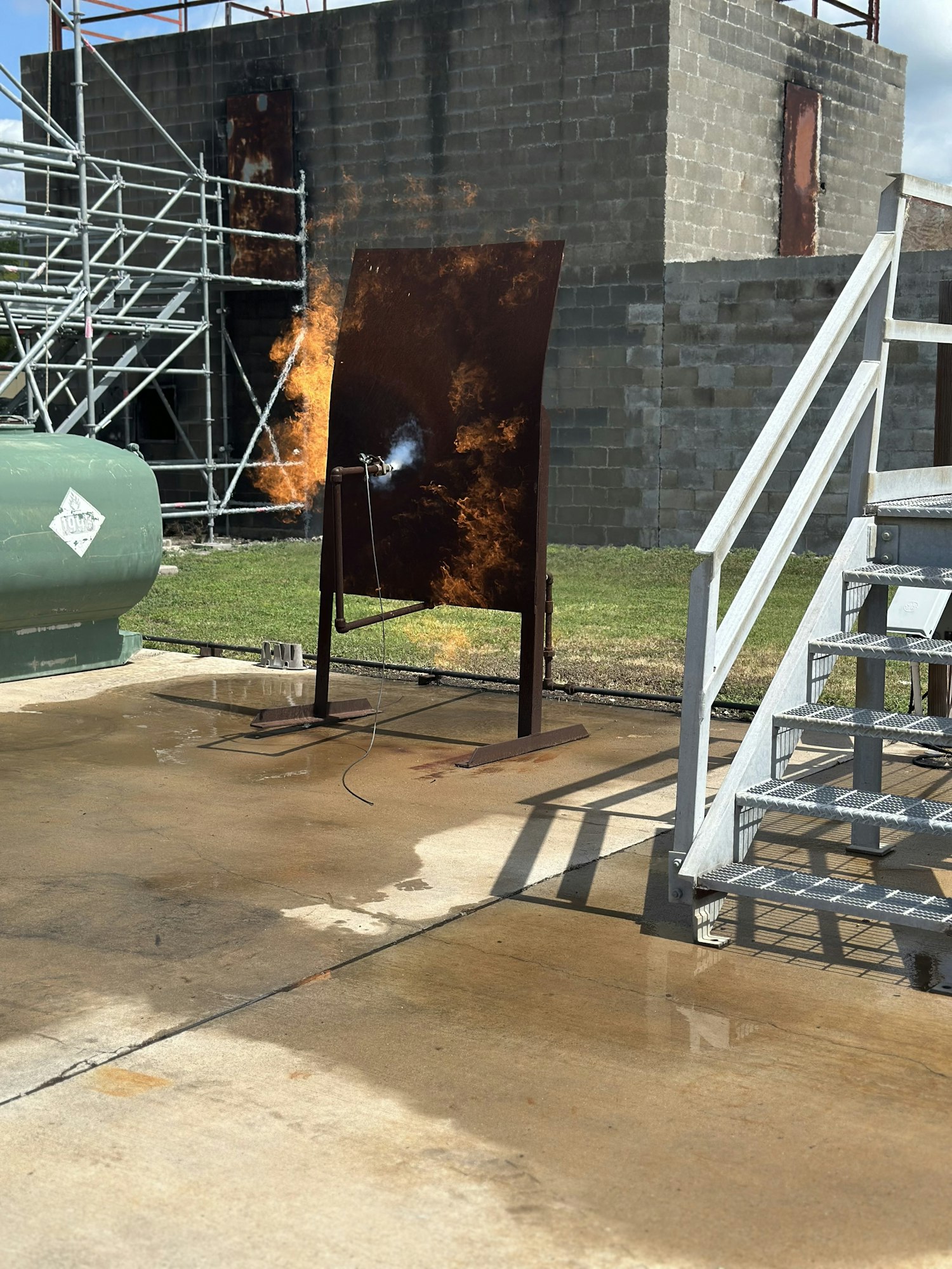 A metal sheet is on fire near a concrete structure, with a green tank and scaffolding in the background.