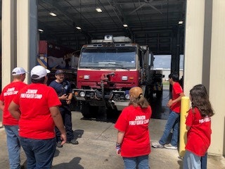 A group in red "Junior Firefighter Camp" shirts stands near a fire truck inside a garage.