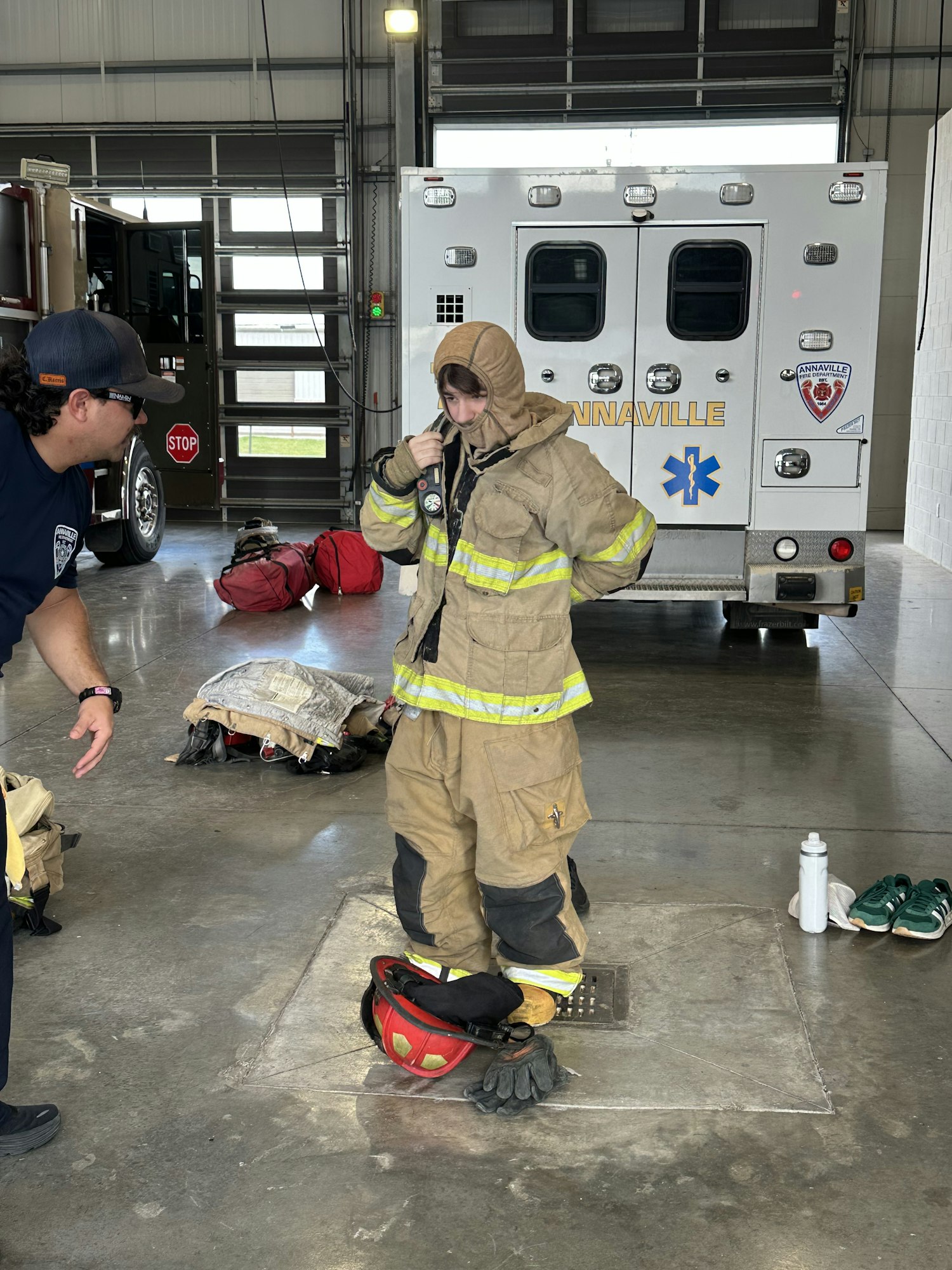 A person is standing in firefighting gear next to a fire engine inside a garage. Another person is nearby.