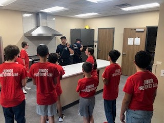 A group of kids in red "Junior Firefighter Camp" shirts listen to an adult in a kitchen setting.