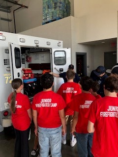 A group of people in red "Junior Firefighter Camp" shirts are gathered around an ambulance, looking inside.