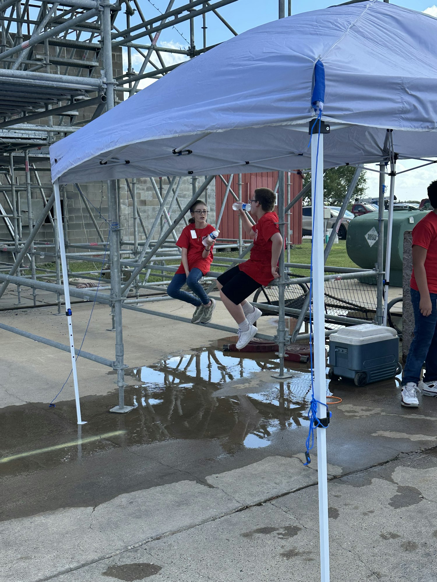 People in red shirts under a canopy next to a puddle on the pavement, with some scaffolding in the background.