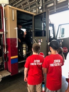 Children wearing "Junior Firefighter" shirts are talking to a firefighter inside a fire truck.