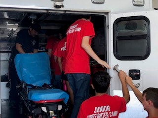 Kids in red shirts labeled "Junior Firefighter Camp" exploring inside an ambulance with a blue stretcher.