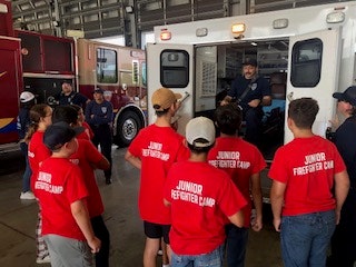 Kids in "Junior Firefighter Camp" shirts listen to a firefighter inside a fire station.
