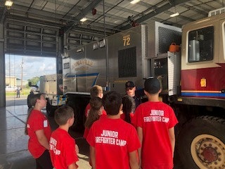 Kids in "Junior Firefighter Camp" shirts listening to an adult in a fire station with fire trucks.