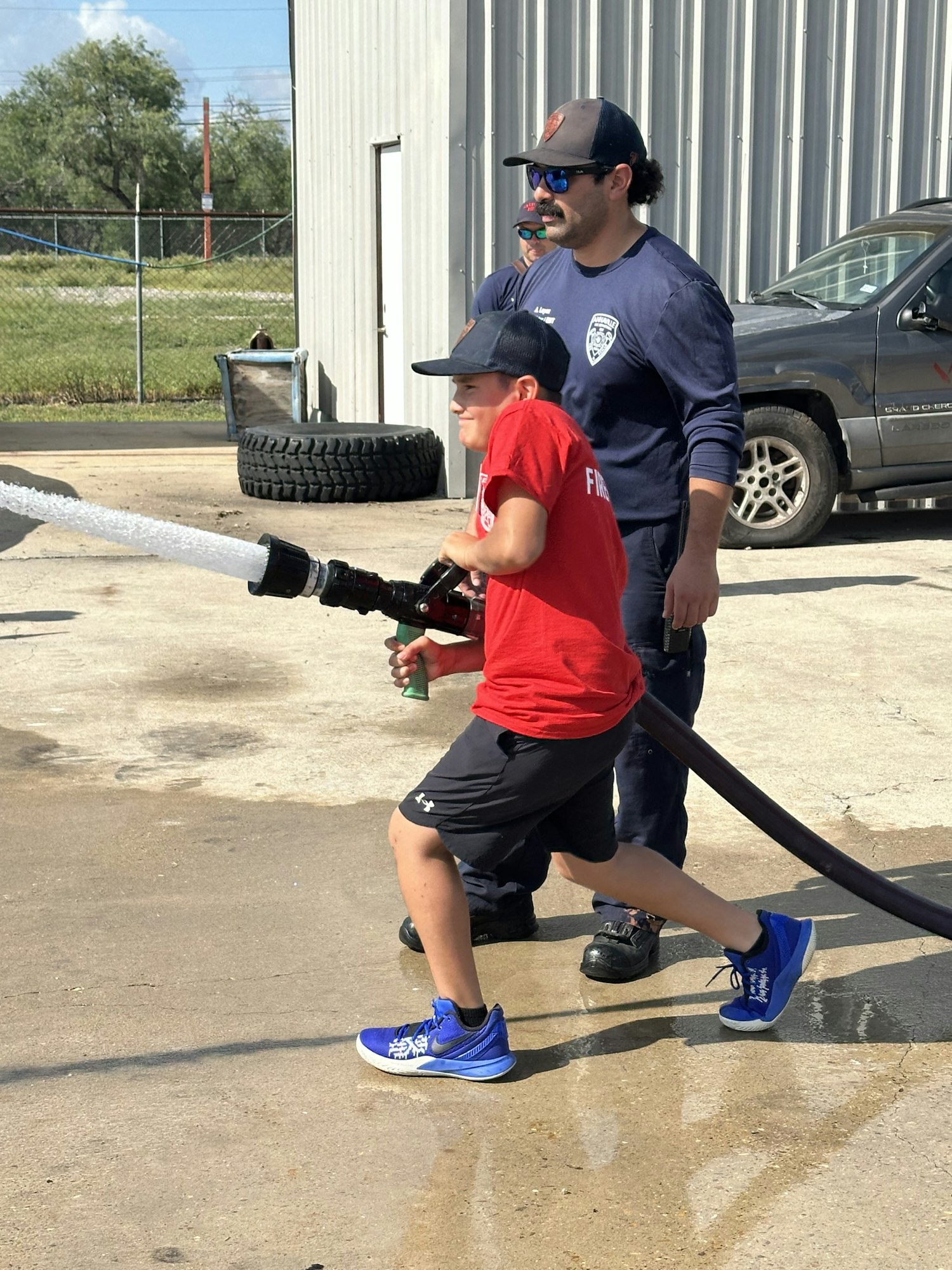 A child sprays water from a fire hose guided by an adult in a uniform, outdoors near a building.