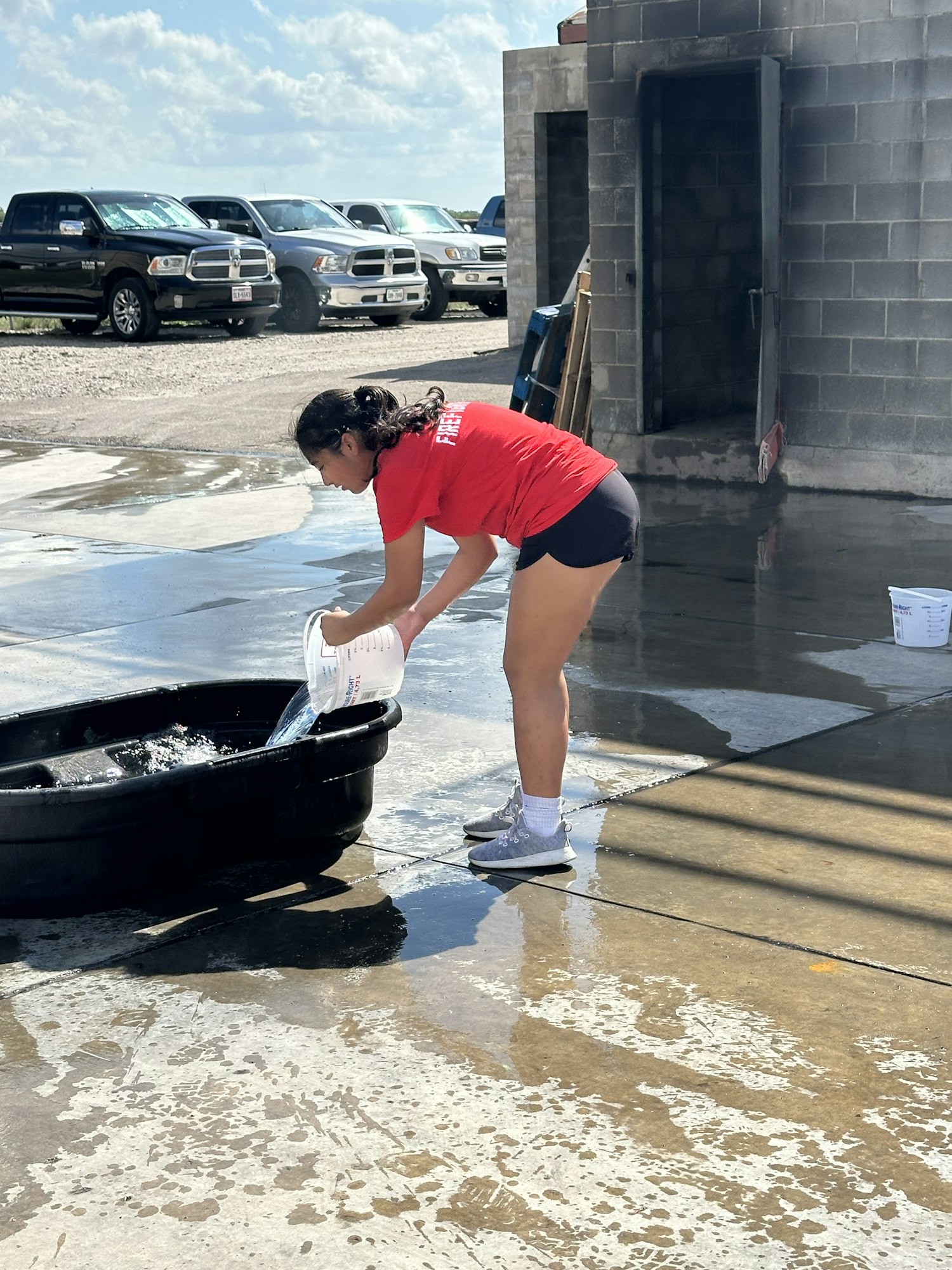 A person in a red shirt pours water from a jug into a black container, surrounded by wet pavement and parked vehicles.