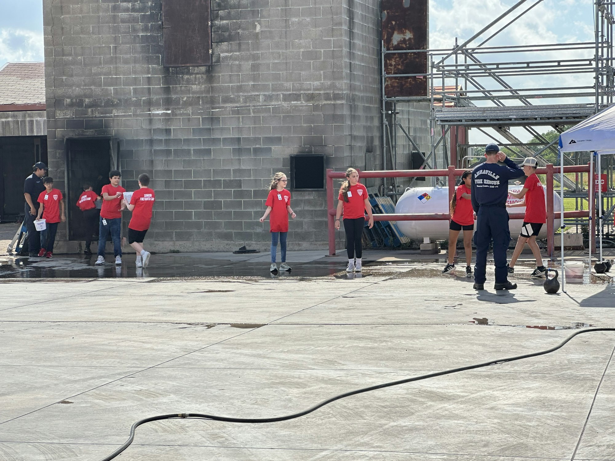 A group of people in red shirts are gathered near a gray building and scaffolding outside.