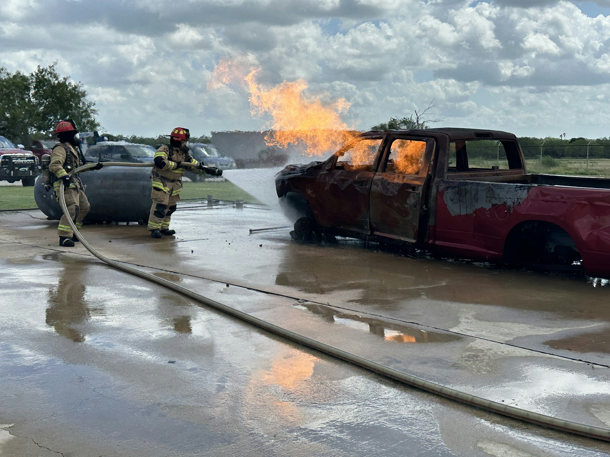 Firefighters extinguishing a burning truck with water hoses.