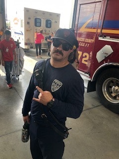 A person in fire department uniform with a walkie-talkie, standing next to a fire truck and ambulance, indoors.