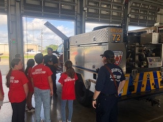 Children in red shirts and two firefighters are gathered around a fire truck in a garage.