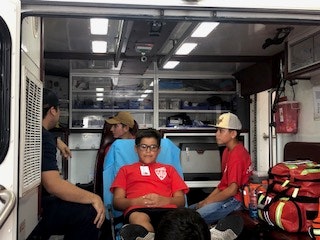 Several people inside an ambulance, one seated on a stretcher, others in seats, with medical equipment visible.