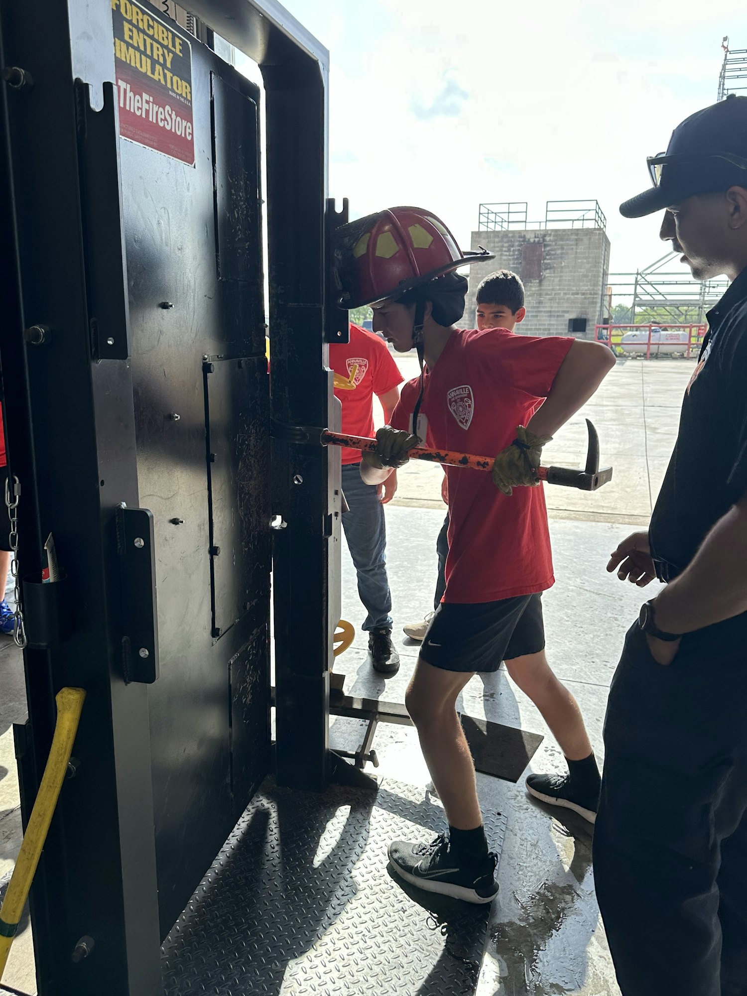 Group of people in red shirts and helmets using a forcible entry simulator.