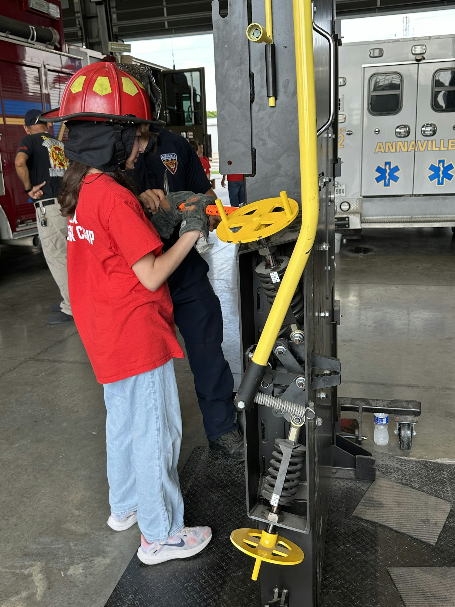 A child in a red shirt interacts with a firefighter at a fire station, with an ambulance visible in the background.