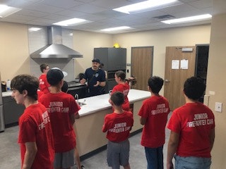 A group of kids in red "Junior Firefighter Camp" shirts listen to an instructor in a kitchen setting.
