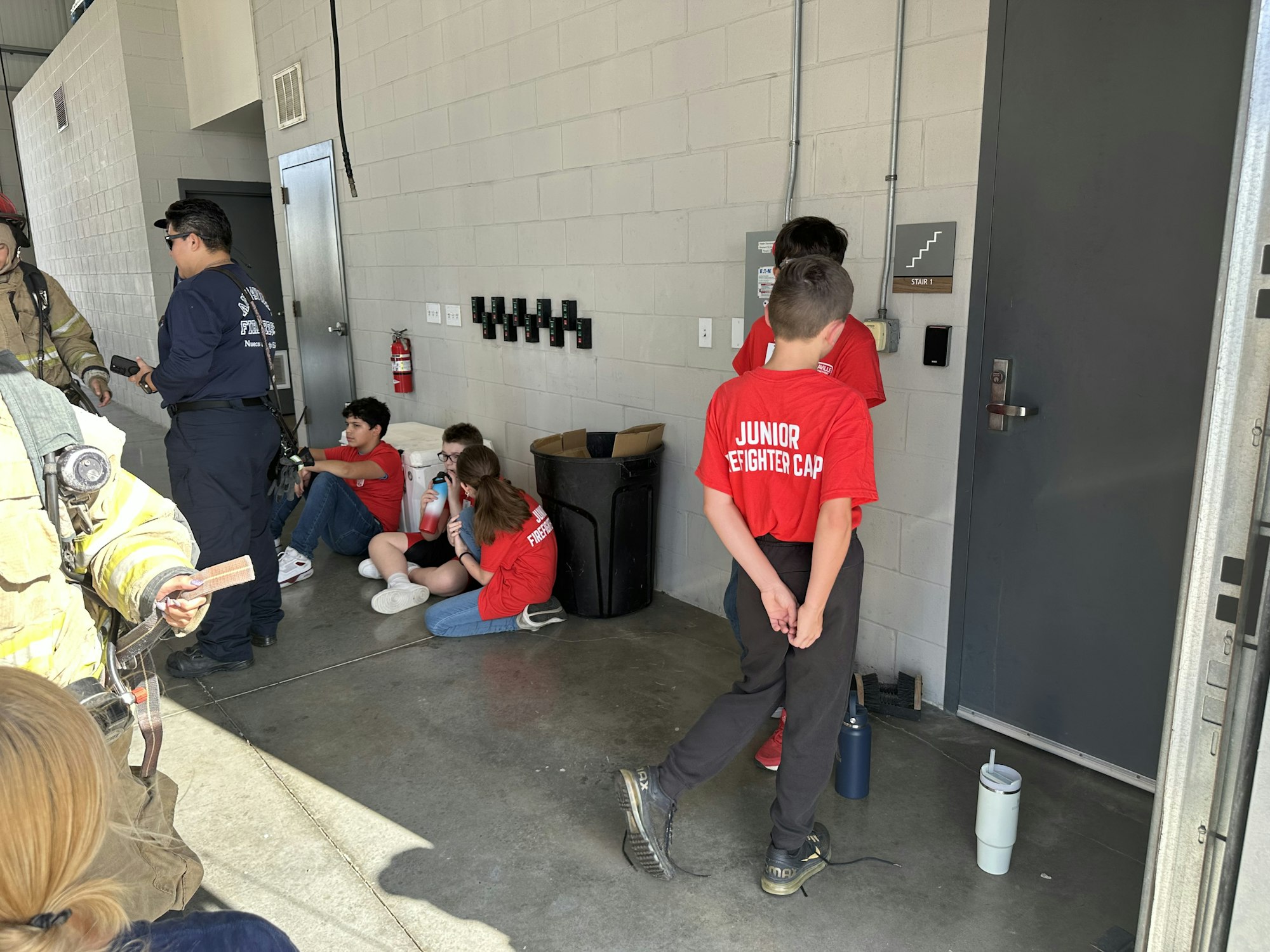 Children in red "Junior Firefighter Camp" shirts and adults, likely firefighters, inside a building, some sitting and some standing.