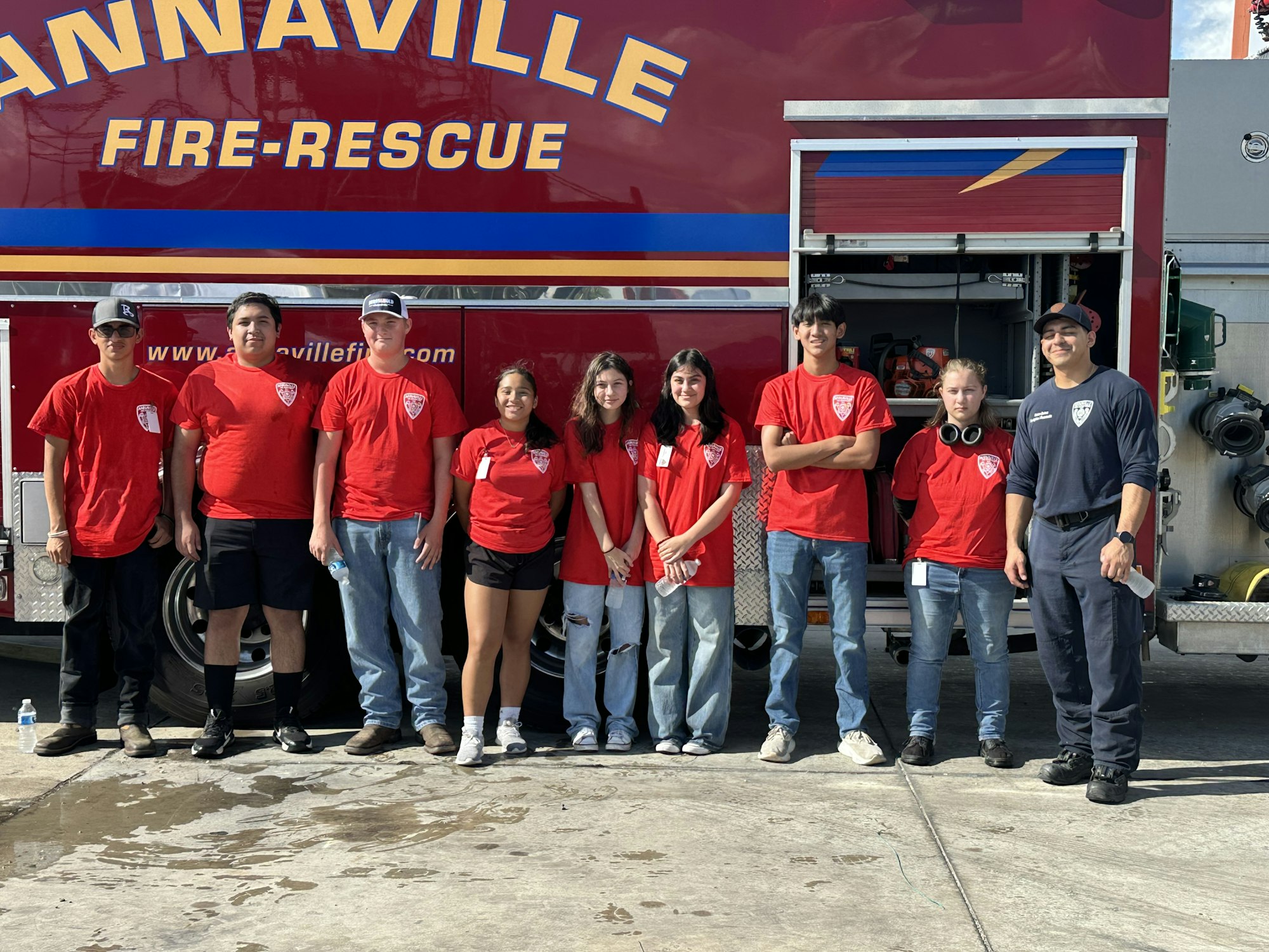 A group of people in red shirts stand in front of a fire-rescue vehicle.