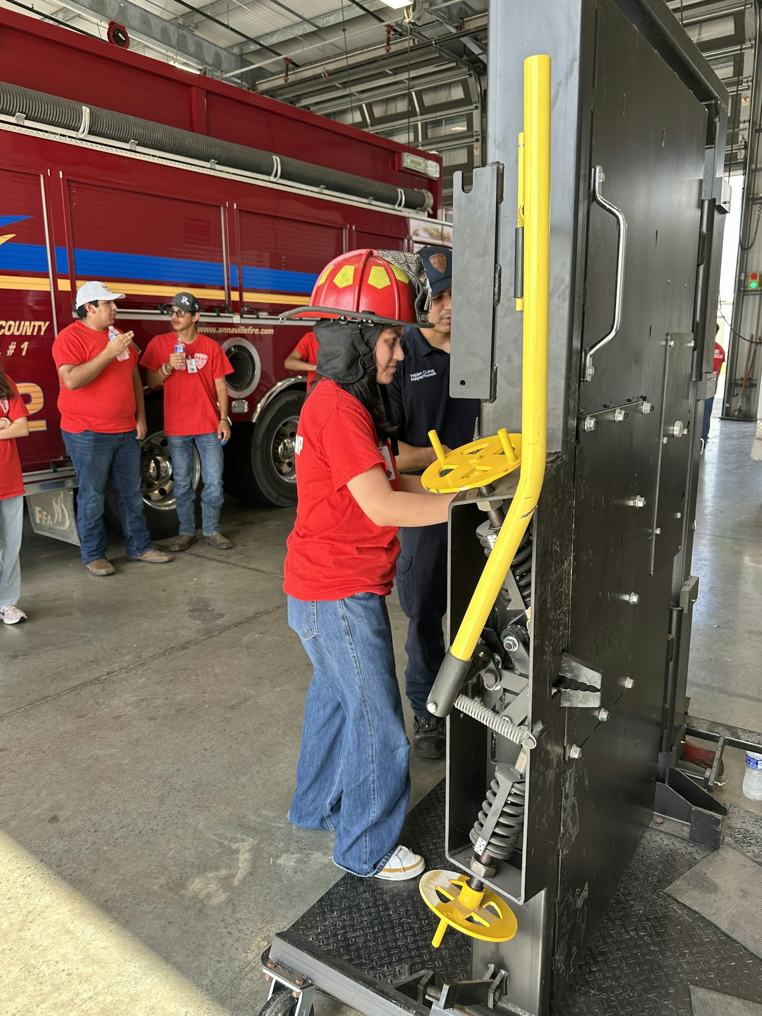 A firefighter training exercise with a group of people, one wearing a helmet, interacting with equipment near a fire truck.