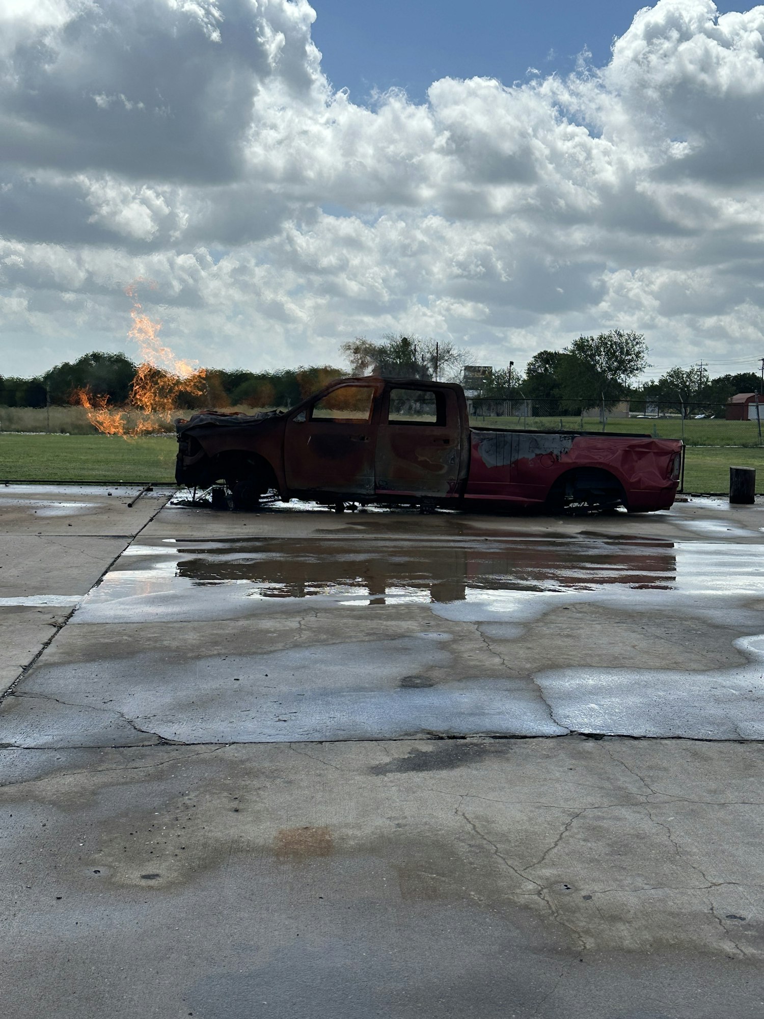 A burnt-out car on a concrete surface with fire and smoke in the background.